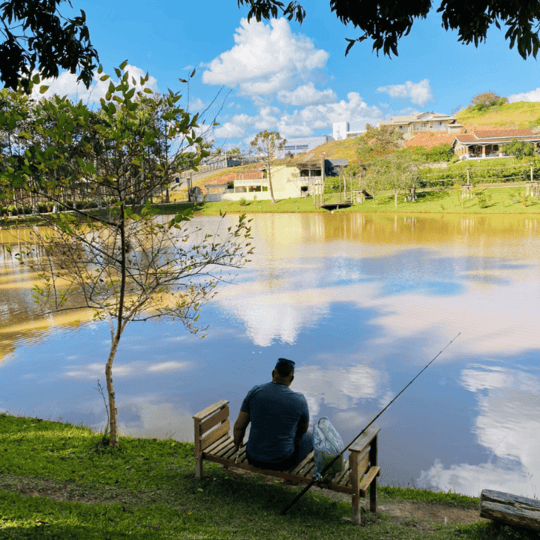 Homem sentado sozinho em uma praça j£ ao lado de um rio tranquilo, cercado por árvores e casas, em um dia ensolarado e de céu limpo, ideal para relaxar e pescar.