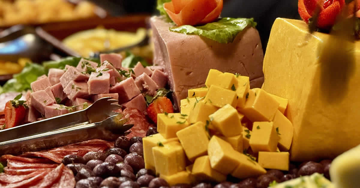 Mesa de jantar com tábua de queijos e frios variados, acompanhada de taças de vinho e pães.
