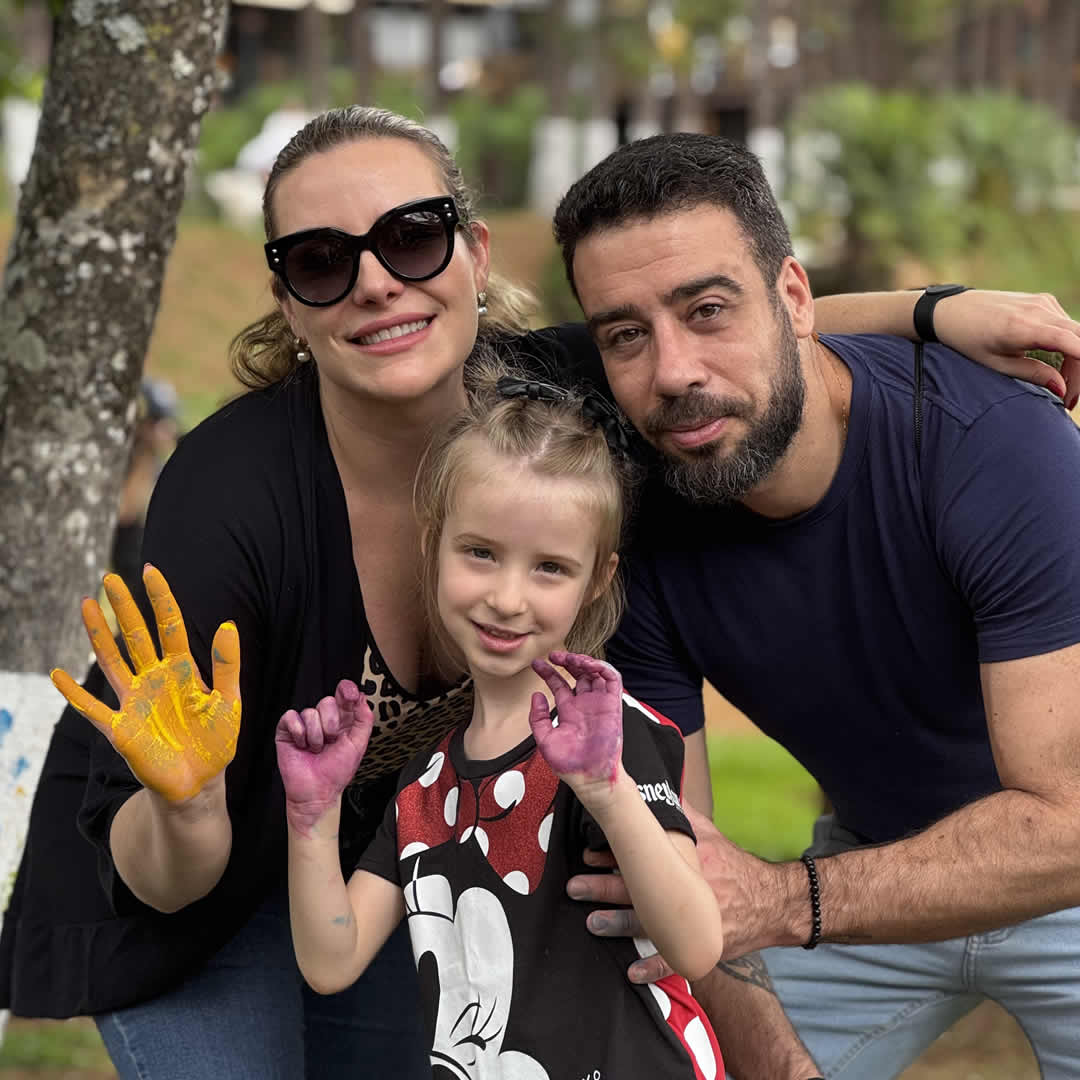 Pais e filha sorrindo com as mãos pintadas, participando de atividade artística juntos