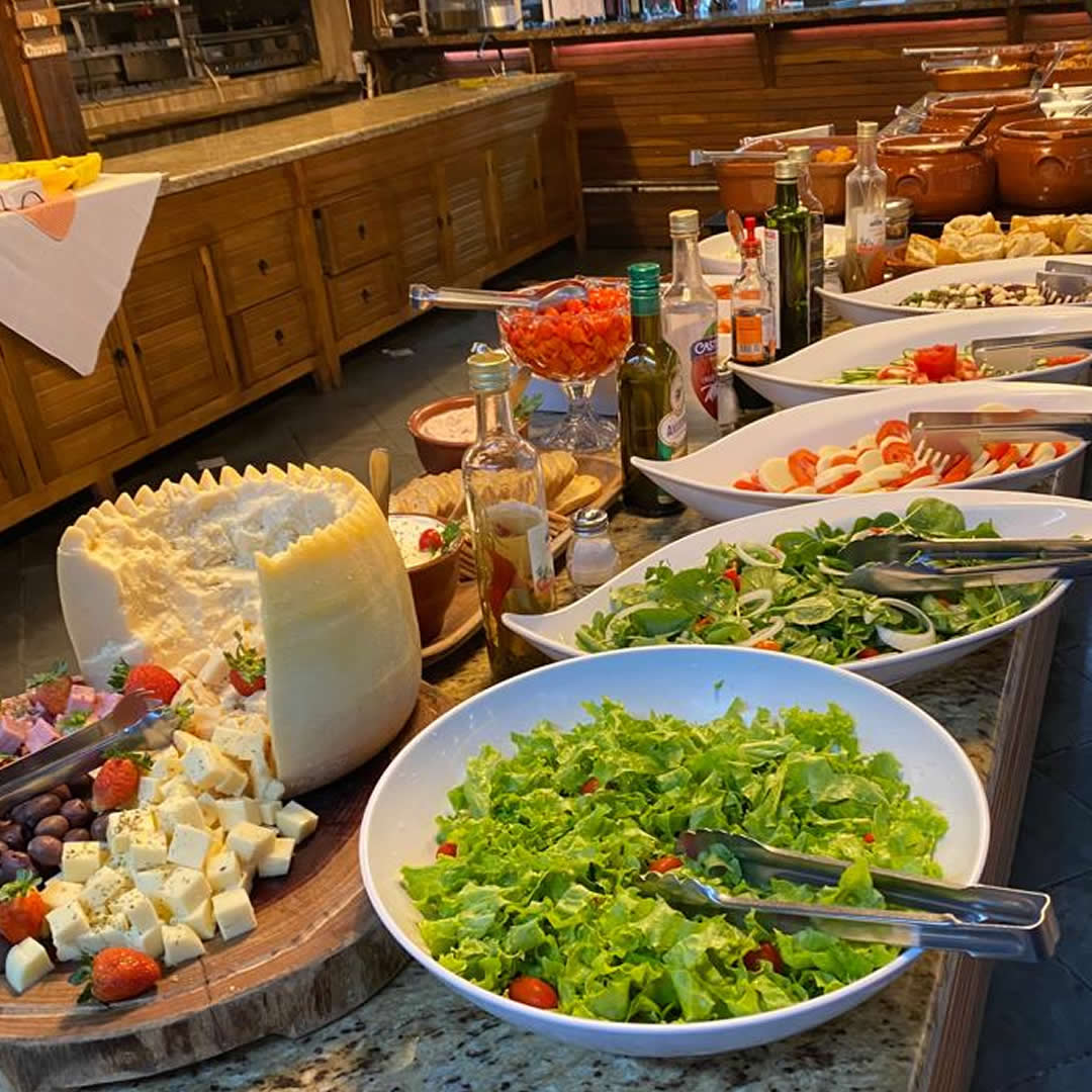 Mesa de jantar com tábua de queijos e frios variados, acompanhada de taças de vinho e pães.