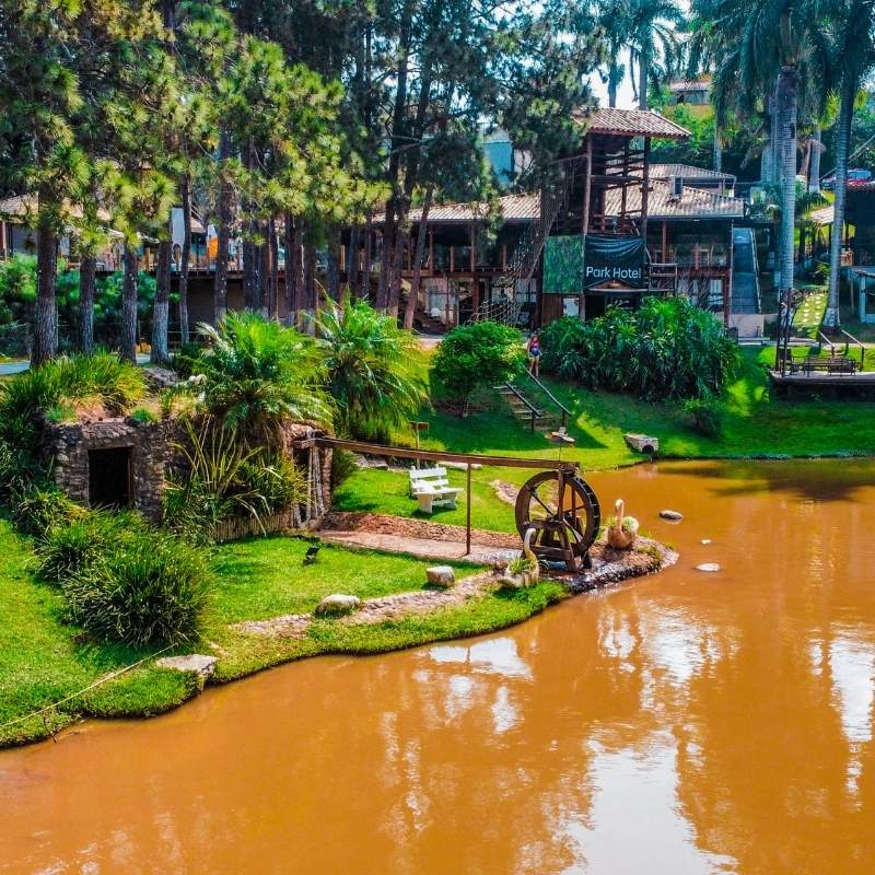 Cenário de parque com lago de água marrom, árvores verdes, roda d'água e estrutura de madeira ao fundo, ideal para lazer e contato com a natureza